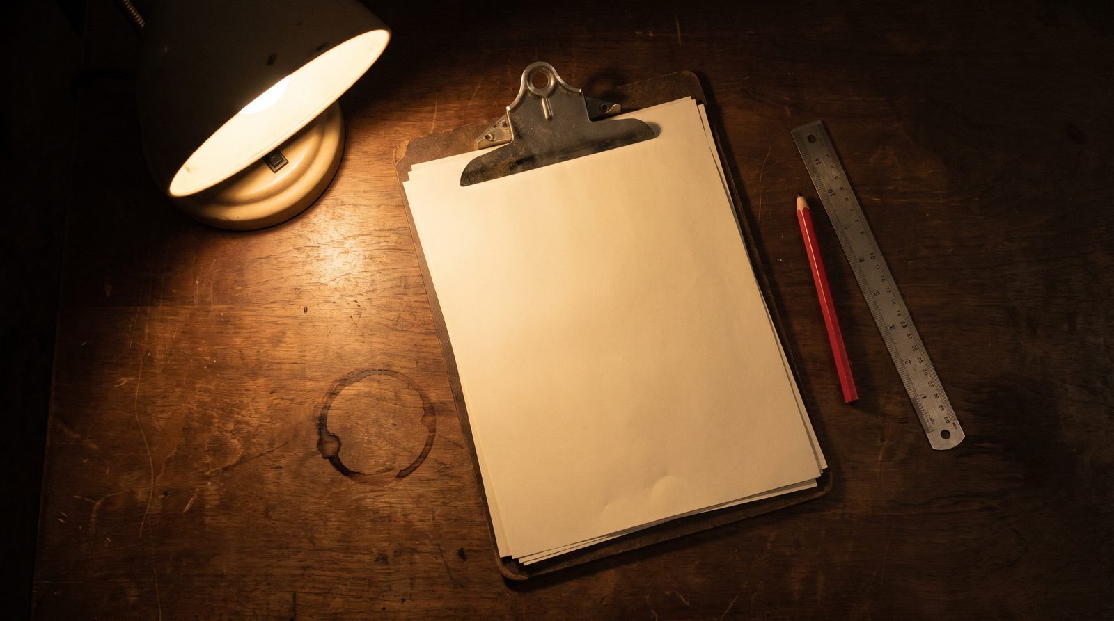 A clipboard holding a blank carbon-copy form pad, a red pencil and a steel ruler on a dark desk under one lamp.